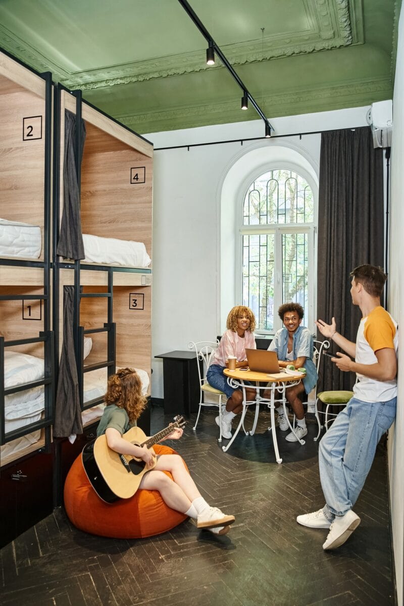 multiethnic friends with laptop and guitar talking in cozy room of modern students hostel, leisure