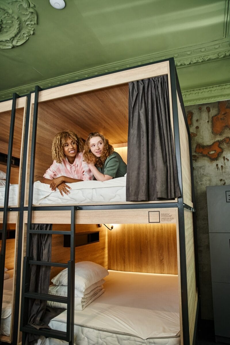 young excited female friends lying on double-decker bed and looking away in modern students hostel
